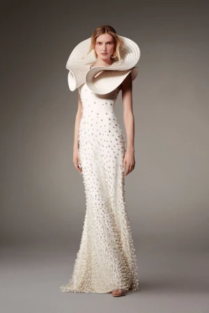 Elegant woman in a white gown with pearl embellishments and a large, textured hat posing in a photo studio with a neutral background.