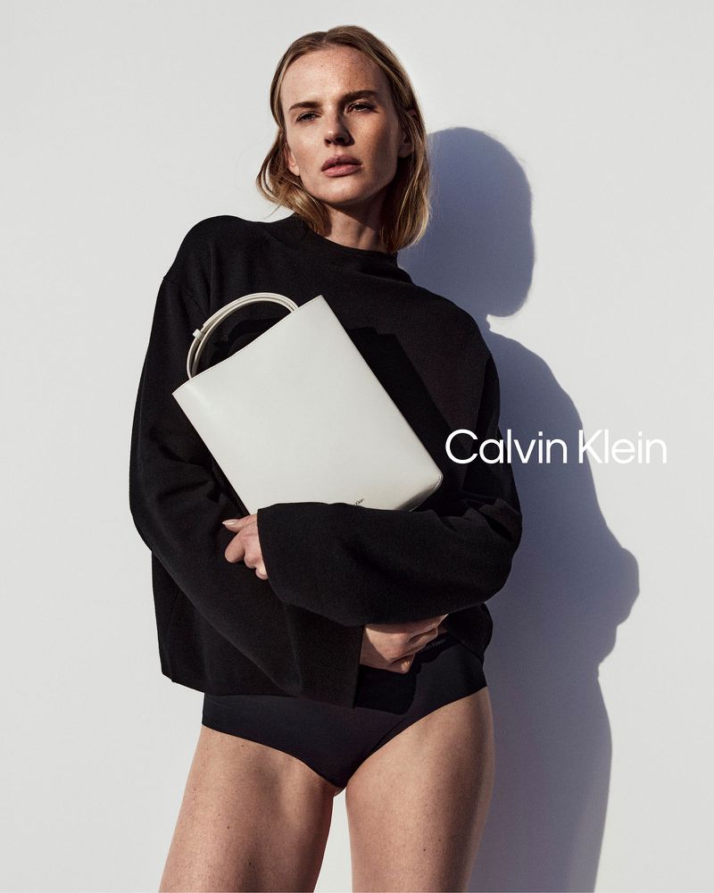 Fashion shot of a young adult woman posing in a black sweater and briefs, holding a white tote bag by Calvin Klein against white wall.