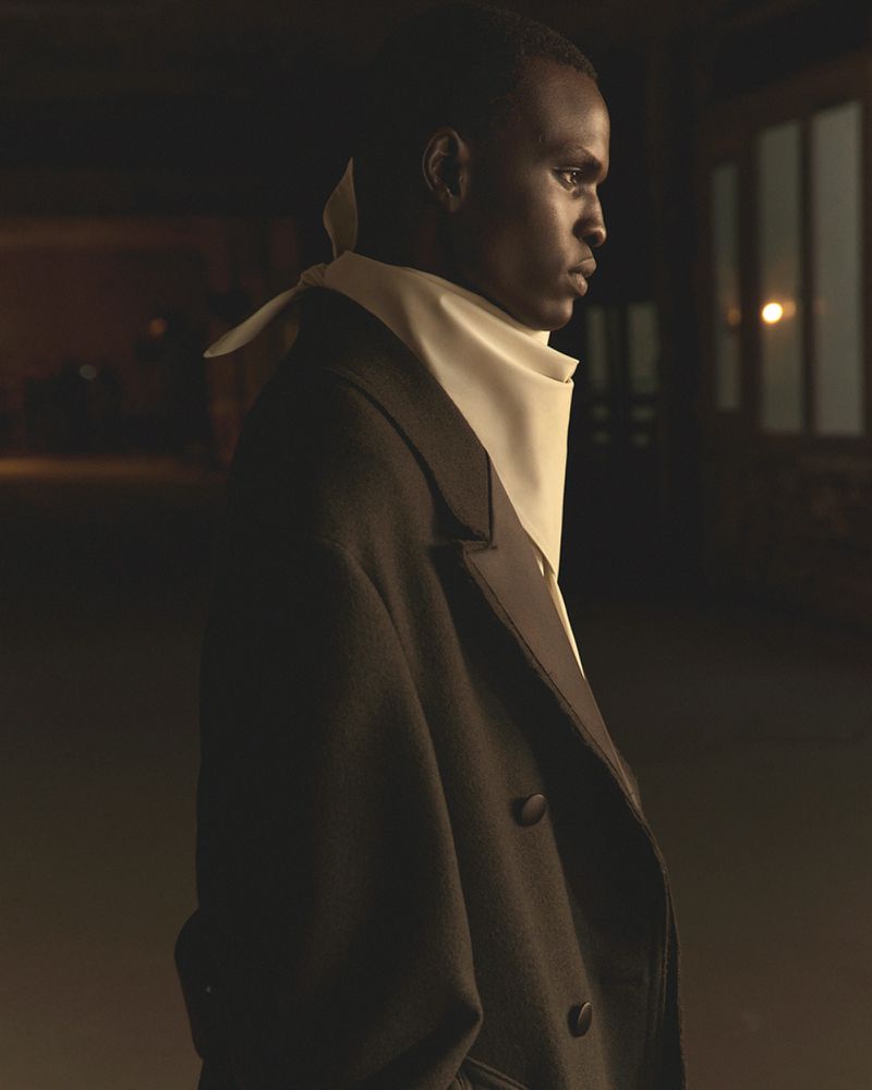 Portrait of a dark-skinned man in a formal black coat and ascot against a dimly lit urban interior background with a window and soft lighting.