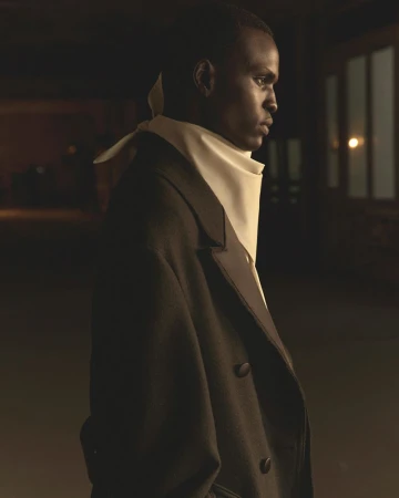 Portrait of a dark-skinned man in a formal black coat and ascot against a dimly lit urban interior background with a window and soft lighting.