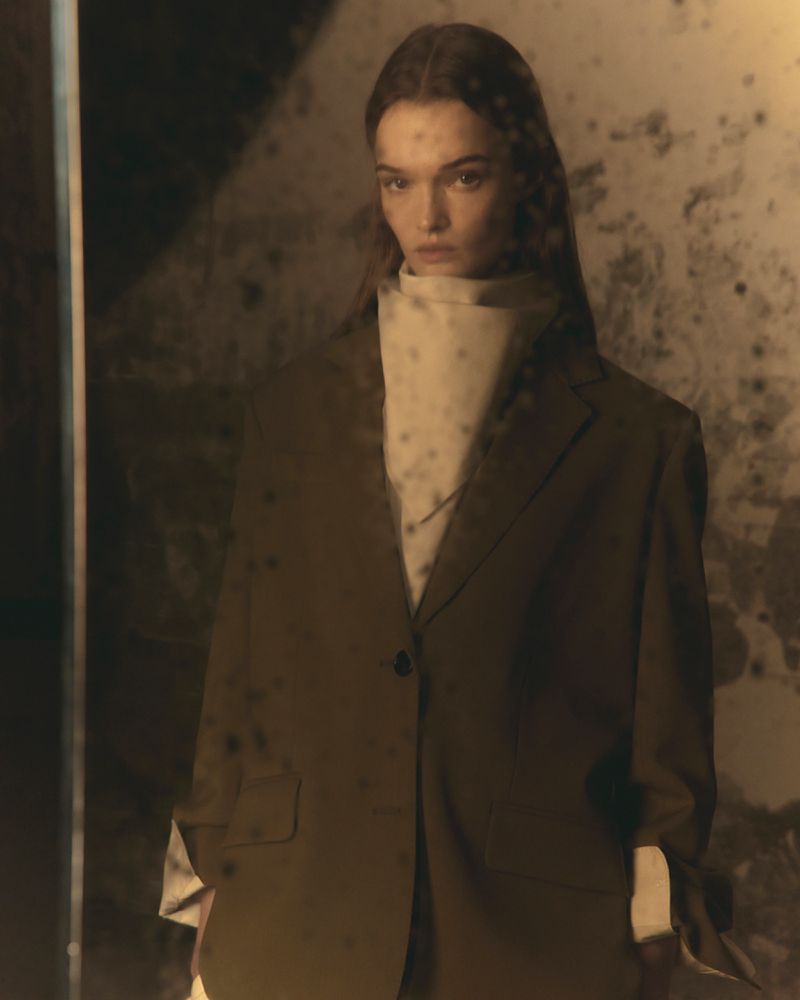 An adult female fashion model poses indoors wearing an oversized olive-green blazer, standing in front of a textured reflective surface.