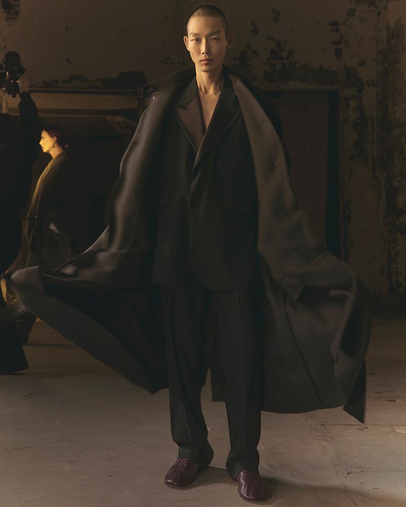 An adult male model with a shaved head is wearing a black suit and coat in a dimly lit room, showcasing modern fashion apparel.