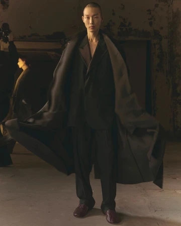 An adult male model with a shaved head is wearing a black suit and coat in a dimly lit room, showcasing modern fashion apparel.