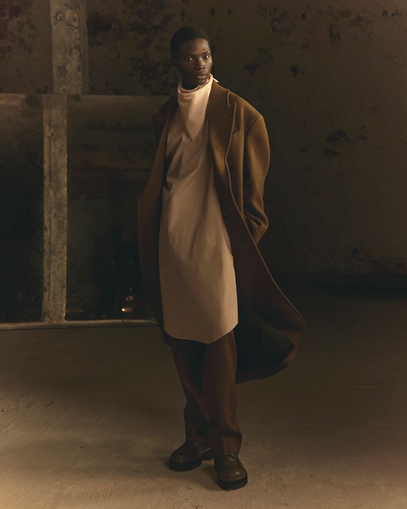 Model in a long brown coat and light beige tunic standing indoors against a weathered concrete pillar background.