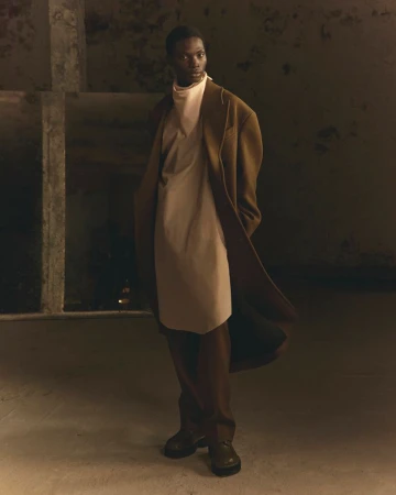 Model in a long brown coat and light beige tunic standing indoors against a weathered concrete pillar background.