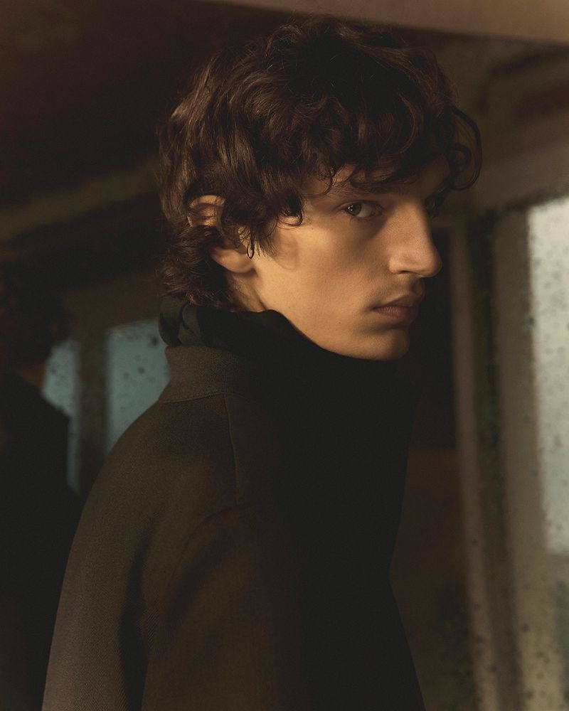 Portrait of a young adult male with dark curly hair wearing a dark coat. The lighting is moody and the background is blurred.