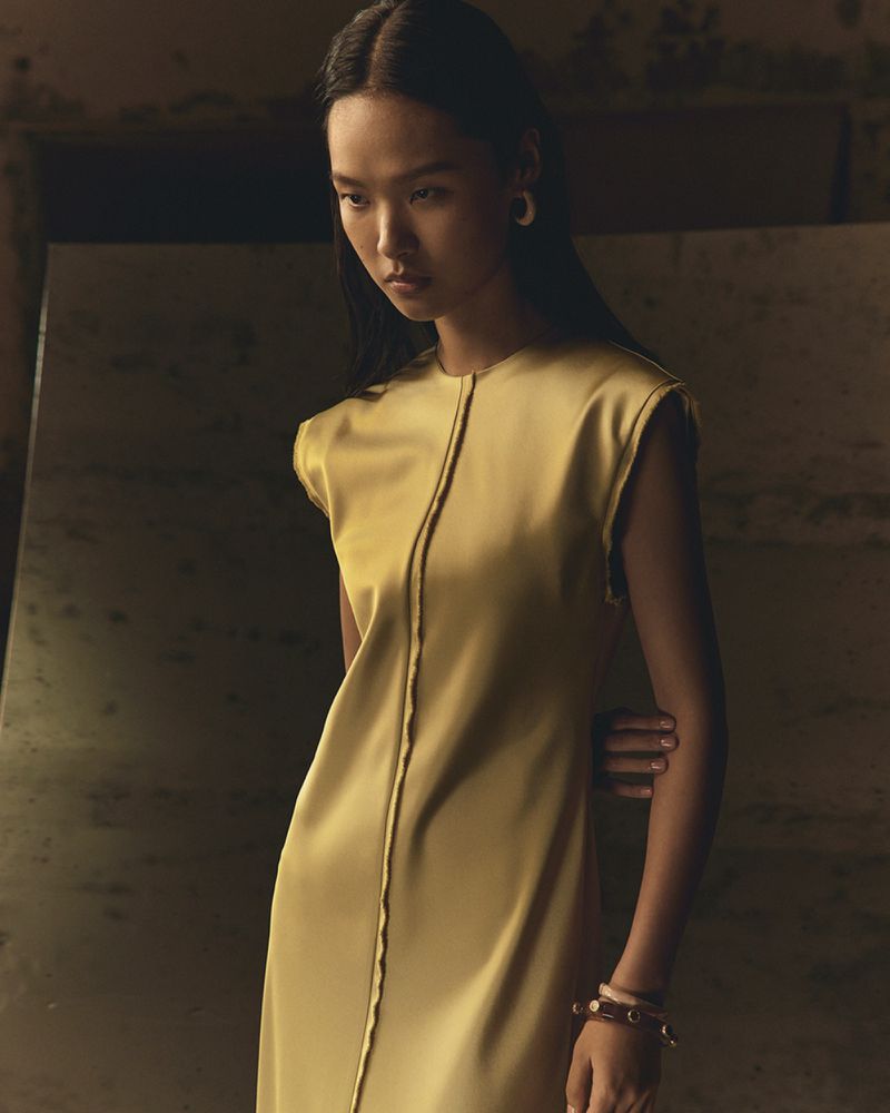 Elegant Asian woman poses in a sleek gold dress with a minimalist design. Soft, diffused lighting highlights the dress's texture.