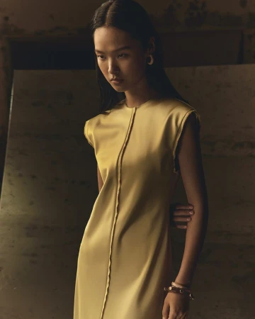 Elegant Asian woman poses in a sleek gold dress with a minimalist design. Soft, diffused lighting highlights the dress's texture.