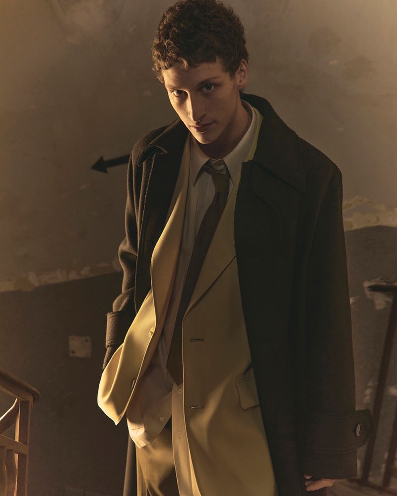 An adult male wearing an overcoat, jacket, shirt, and tie stands inside a building looking down at the camera in a medium shot.