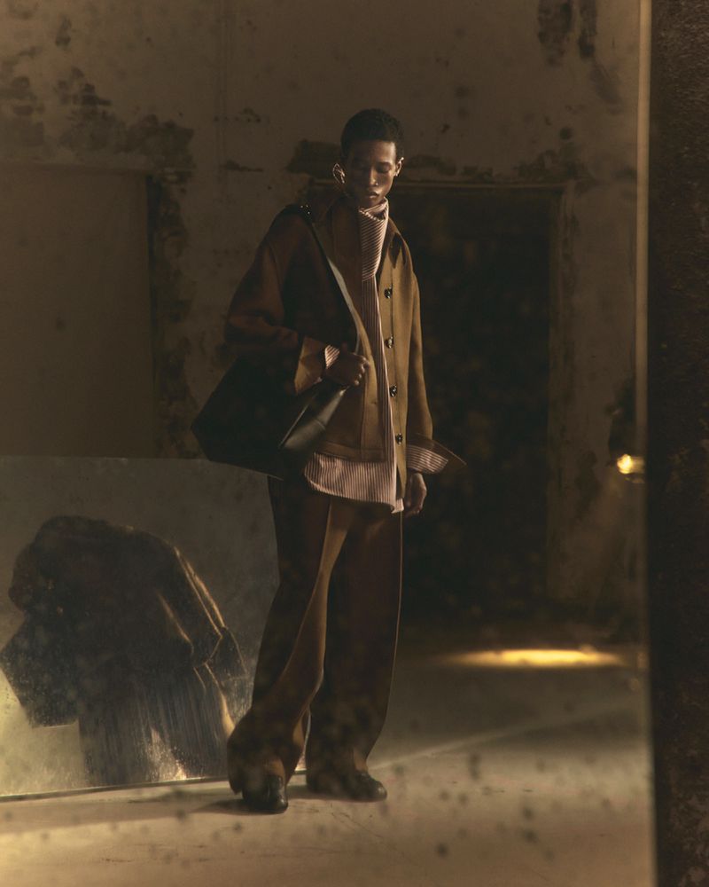 A male fashion model poses in an open brown coat, wide brown pants, and carrying a dark shoulder bag, inside an aged interior space.