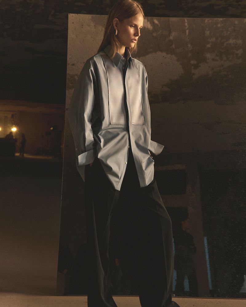 Fashion photo of a slender woman in a collared top with wide, loose-fitting dark pants, posing in a dimly lit, industrial environment.