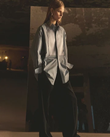 Fashion photo of a slender woman in a collared top with wide, loose-fitting dark pants, posing in a dimly lit, industrial environment.
