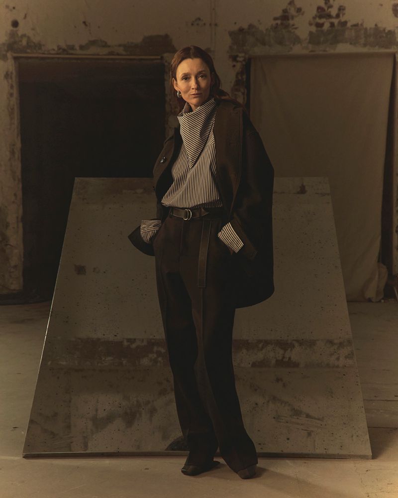 Fashionable adult woman in a black coat and wide-legged trousers stands indoors, posing next to a large mirror in a minimalist style.