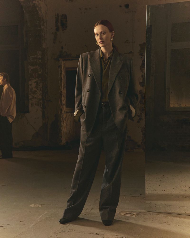 Adult female fashion model in a grey oversized suit stands in a dimly lit interior space near a mirror with a second person in background.