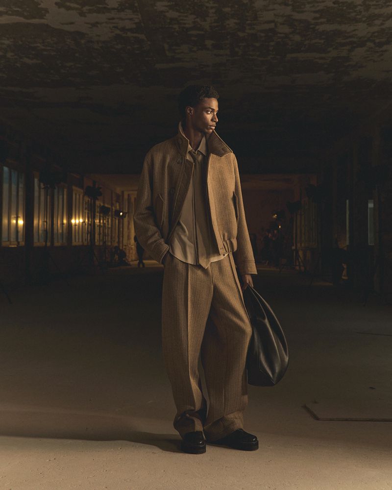A stylish Black male model poses in a tan jacket and pants with a bag in an urban setting under dramatic lighting, looking off-camera.