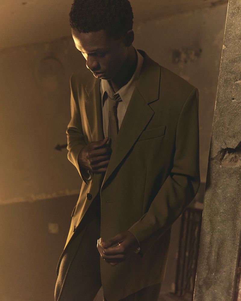 Portrait of a young African-descent man wearing an olive green suit, tie, and dress shirt in an indoor, dimly-lit location with concrete walls.