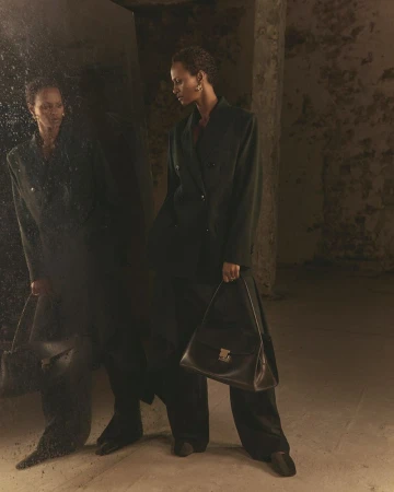 Model wearing a dark blazer and trousers holding a structured leather handbag next to a speckled mirror in an interior space