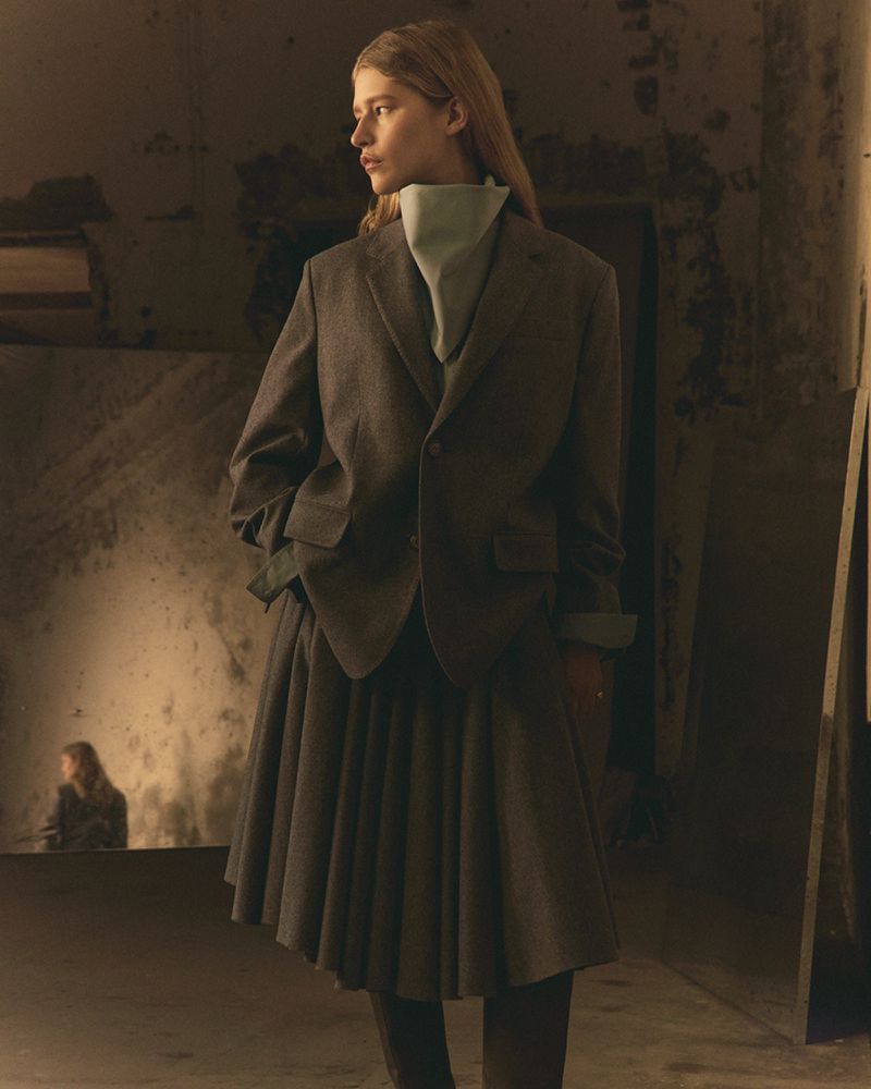 Fashionable adult female model in a blazer and skirt posing in a dimly lit indoor space with textured walls and light from the side.