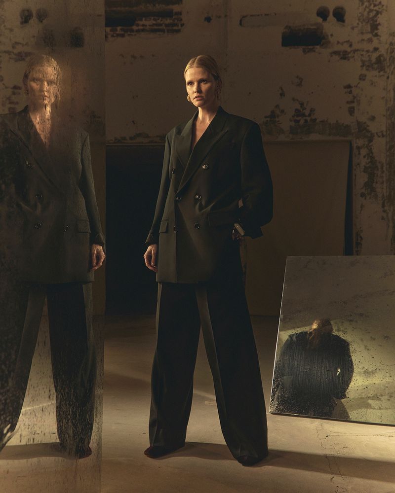 A woman in a dark oversized double-breasted suit poses indoors with a mirror reflecting her image, creating a high-fashion editorial look.