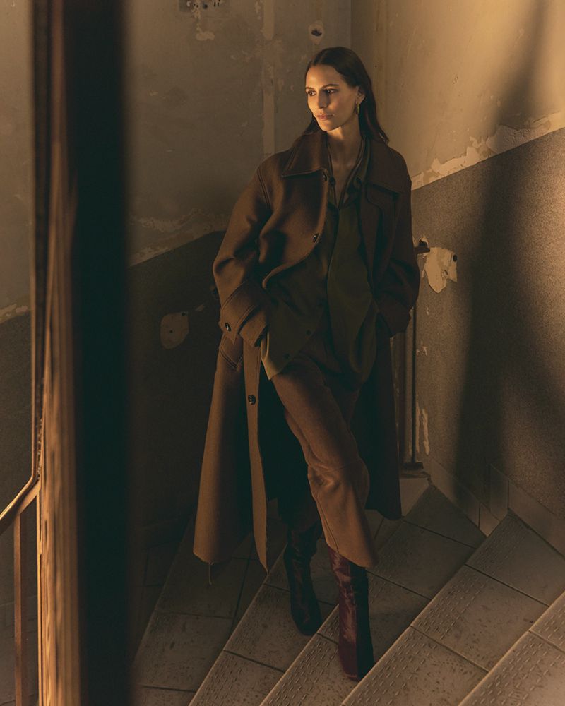 Woman in a brown overcoat and burgundy boots walks down stone stairs in an indoor environment with neutral colors and diffused lighting.