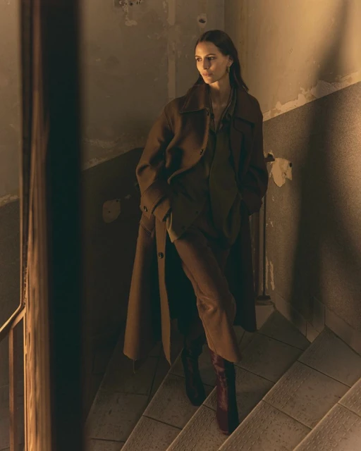 Woman in a brown overcoat and burgundy boots walks down stone stairs in an indoor environment with neutral colors and diffused lighting.