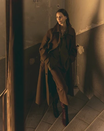 Woman in a brown overcoat and burgundy boots walks down stone stairs in an indoor environment with neutral colors and diffused lighting.
