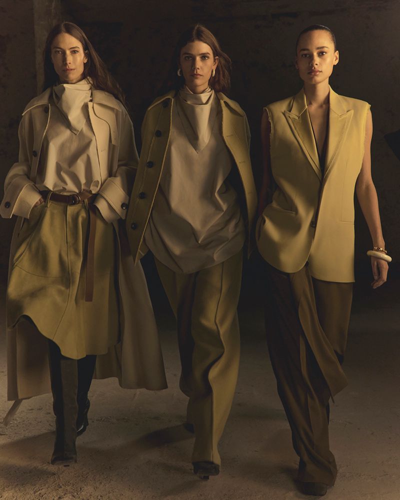 Three women model neutral-toned fashion in studio. Two wear coats, one a vest suit, all in pants, styled in contemporary editorial mood.