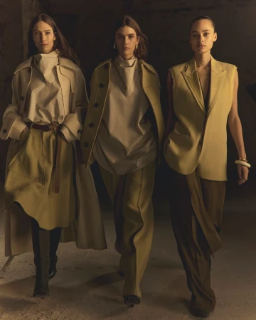Three women model neutral-toned fashion in studio. Two wear coats, one a vest suit, all in pants, styled in contemporary editorial mood.