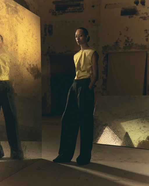 Indoor shot of a young adult woman in a gold sleeveless top and dark wide-leg pants, with a mirror reflection, in a dimly lit room.