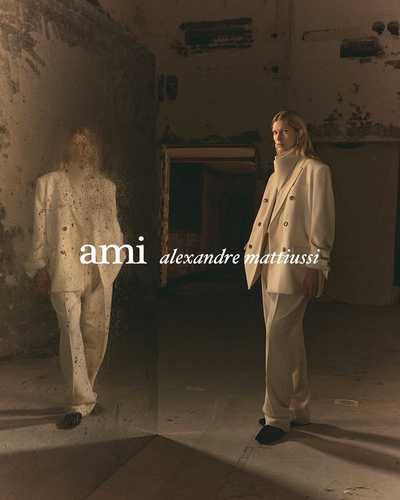 Fashion shot of a blonde woman in a cream-colored suit posing in a dimly lit, grungy interior, with a partial reflection visible.