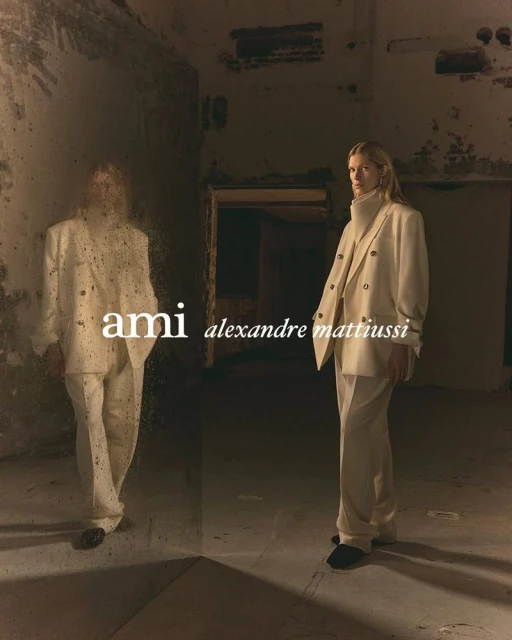 Fashion shot of a blonde woman in a cream-colored suit posing in a dimly lit, grungy interior, with a partial reflection visible.