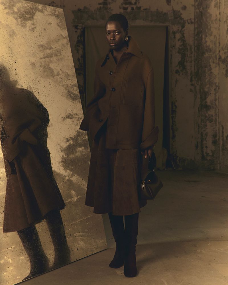 A dark-skinned fashion model poses in a brown dress and jacket with a black handbag against a textured wall in a dimly lit studio.