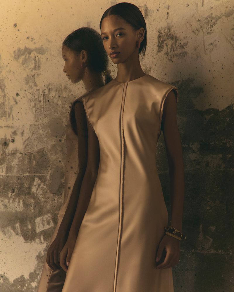 Model wearing a minimalist beige dress standing against a textured concrete wall with a mirror reflection