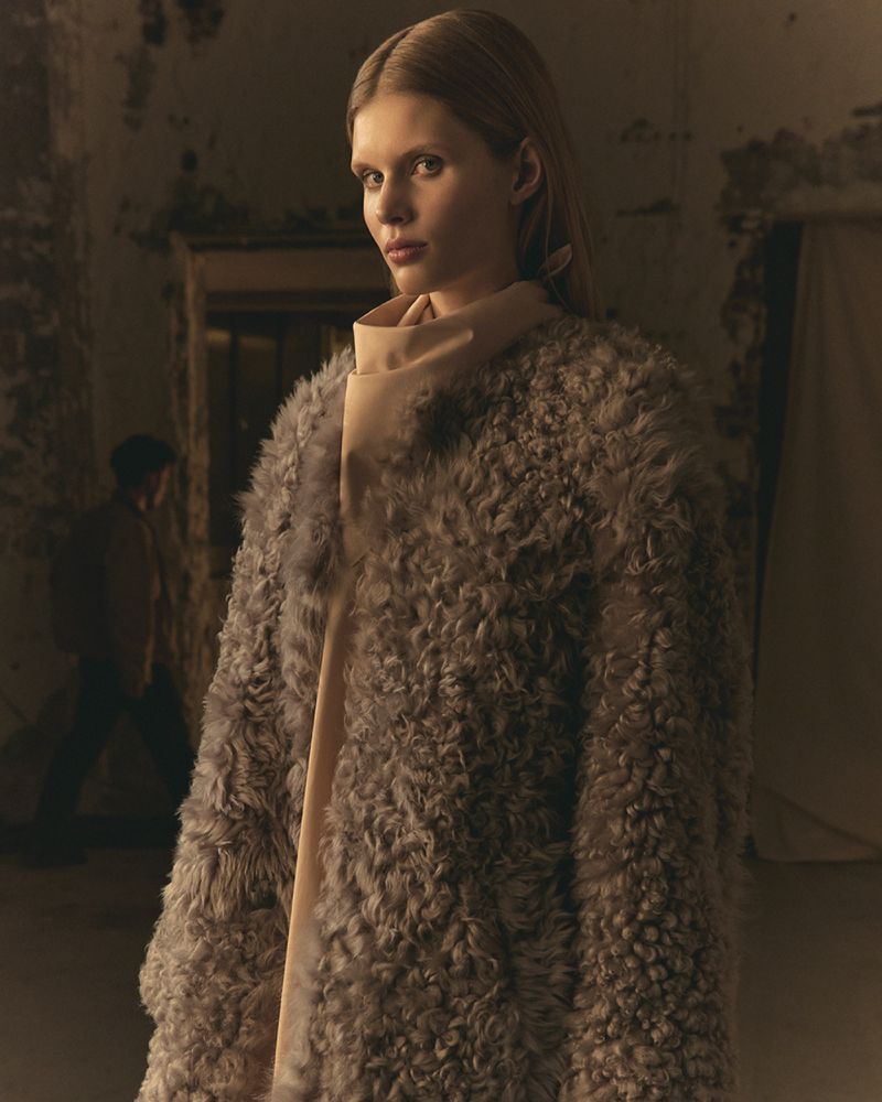 Indoor fashion shot of a young adult female model wearing a fur coat and scarf, looking at the camera, with a person blurred in the background.