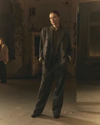 Model standing in a structured grey double-breasted suit and trousers inside a rustic interior space with distressed walls.