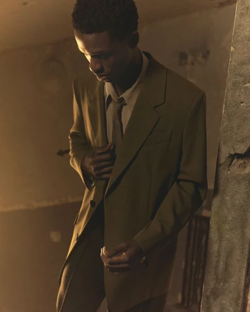 Man wearing an olive suit jacket and matching tie standing indoors in a dimly lit, atmospheric room.