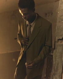 Man wearing an olive suit jacket and matching tie standing indoors in a dimly lit, atmospheric room.