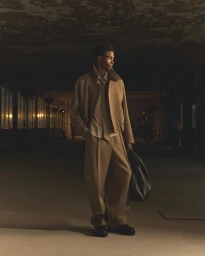 Model standing in a moody interior space wearing a camel jacket and trousers, holding a large black bag
