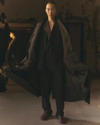 Model with a shaved head wearing a dark blazer, trousers, and long coat for the Ami Fall-Winter 25 collection.