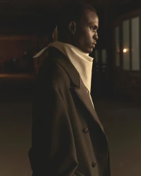 Profile portrait of a male model wearing a dark coat and high white neck scarf in a dimly lit interior.