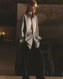 Model standing in a long grey collared shirt and dark trousers against a concrete backdrop under moody lighting.