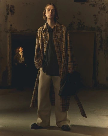 Model wearing a plaid trench coat and loose trousers standing in a dimly lit industrial space.