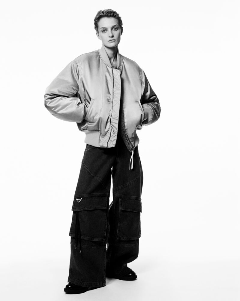 Fashion shot of a woman with short hair in a bomber jacket and wide-leg pants, standing in a studio with a bright background.