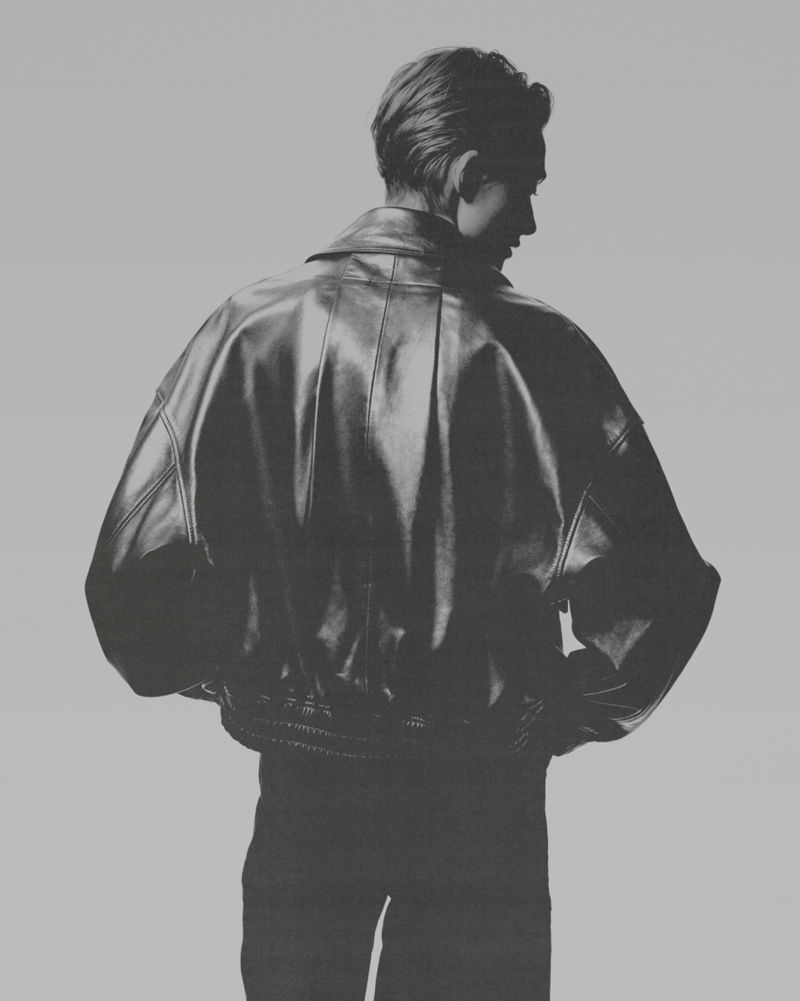 Model seen from behind wearing an oversized black leather jacket against a seamless grey backdrop.