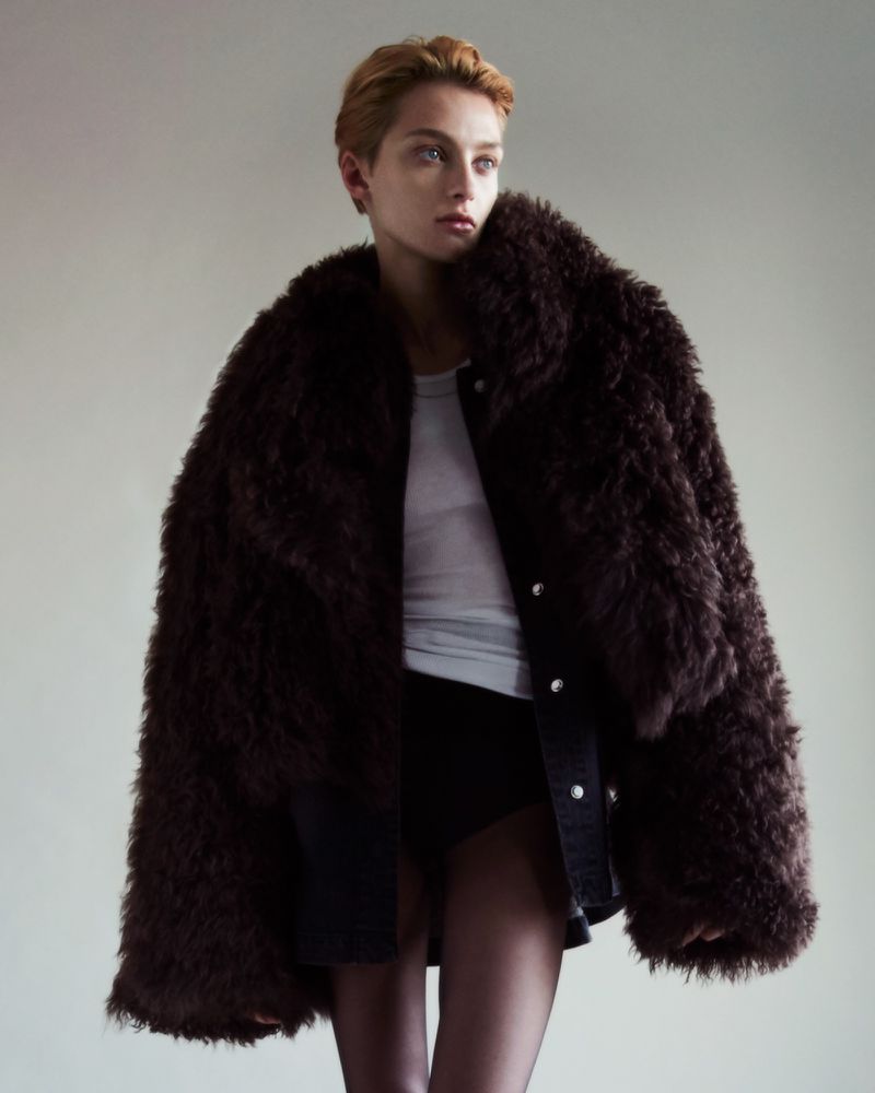 A person in a brown faux fur coat, white tank top, and black shorts is posing against a neutral background in a studio shot.