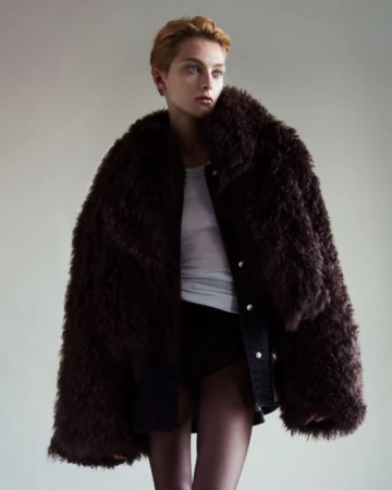 A person in a brown faux fur coat, white tank top, and black shorts is posing against a neutral background in a studio shot.