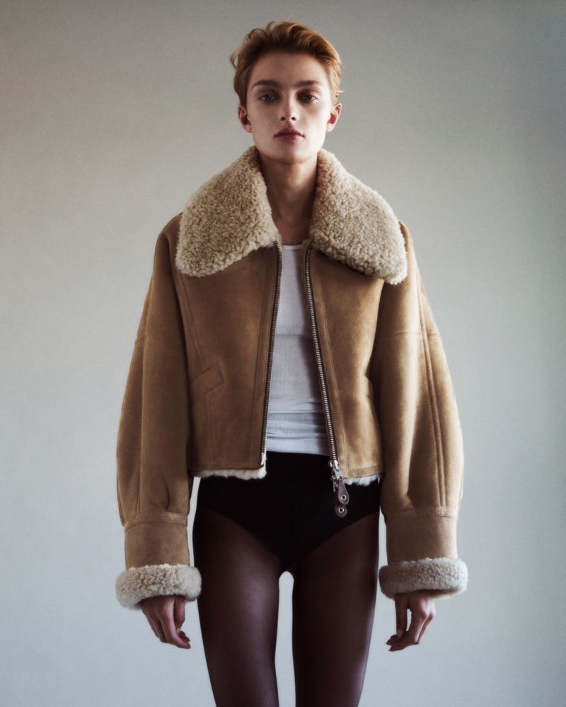 Fashion shot of an adult female in a tan shearling jacket and black bottoms against a neutral background in a photo studio.