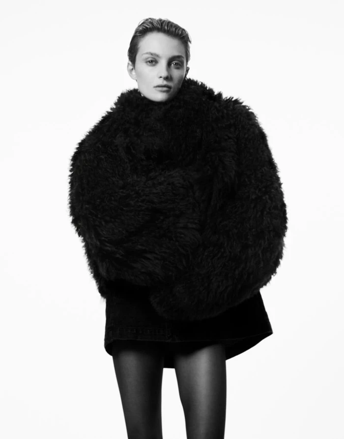 Model with short hair wearing a voluminous black shearling jacket against a white background