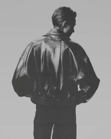 Model seen from behind wearing an oversized black leather jacket against a seamless grey backdrop.