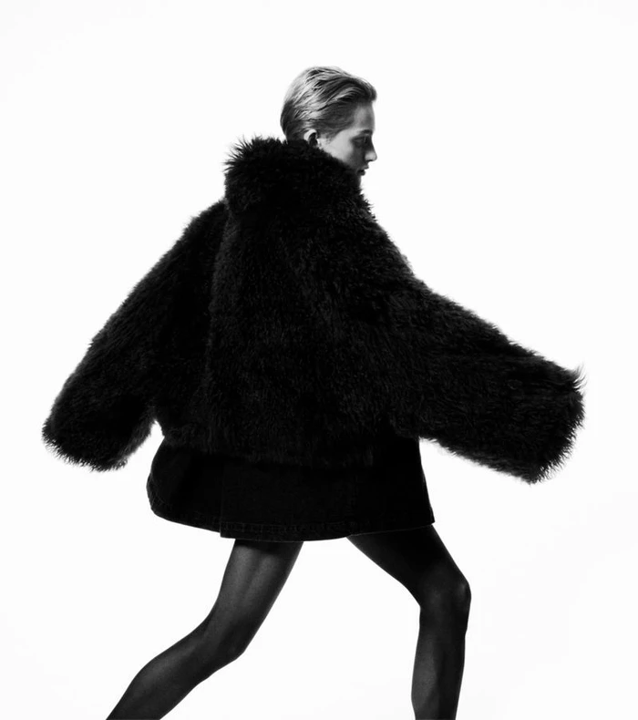 Model in voluminous black furry jacket posing dynamically against a stark white studio background